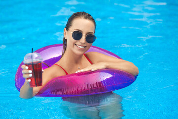 Portrait of young and beautiful smiling woman in sunglasses floats in neon pink swim rubber ring in the pool at aqua park.