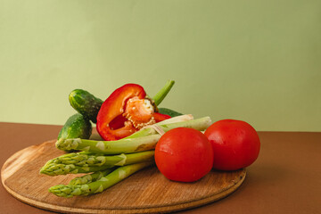 Vegetables lie on a wooden board: tomatoes, asparagus, cucumbers, red bell peppers. brown, light green background. place for text.