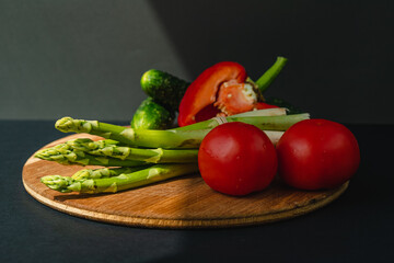 Vegetables lie on a wooden board: tomatoes, asparagus, cucumbers, red bell peppers. brown, dark gray background. place for text.