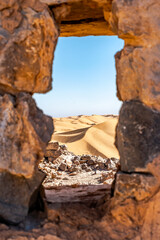 Aerial view through a blurred stone window from the ruines of Djebel Baroun mountain. Taghit, Bechar, Algeria Sahara desert. sand dunes with a blue clear sky. Two far tiny tourists climbing the dune.