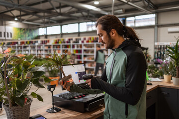 A man behind the cashier stand is scanning a price tag on a potted plant with a barcode reader. - obrazy, fototapety, plakaty
