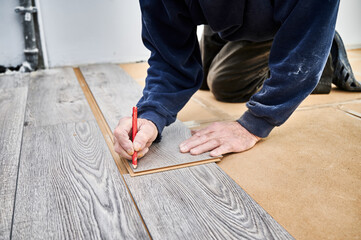 Close up of man construction worker with pencil in hand drawing mark on laminate wooden plank. Male worker preparing laminate boards for floor installation in apartment under renovation.