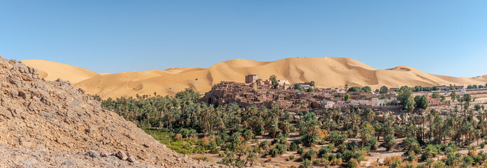 Panoramic view of Taghit in Bechar. Algerian Sahara desert oasis palm trees, trees, sand dunes and ksar old dry stone buildings. Aerial view from rocky mountain Djebel Baroun in foreground.  - obrazy, fototapety, plakaty