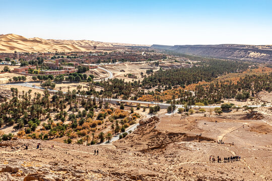 Aerial view from Djebel Baroun rocky mountain in Taghit town of Bechar, Algeria. Far unrecognizable tourists walking in line. Palm trees along the Oued Zouzfana dried river. Sand dunes and blue sky.