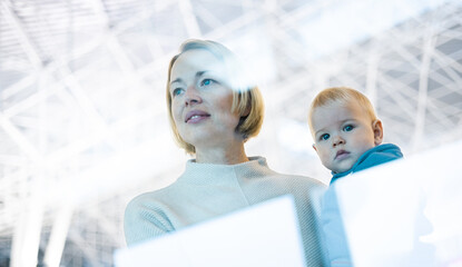 Thoughtful young mother looking trough window holding his infant baby boy child while waiting to...