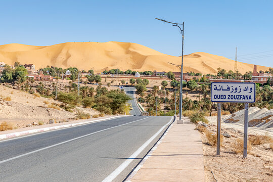 Oued Zouzfana road panel in arabic and french language. Empty road with light poles and dry stones in the roadside, palm trees and trees with buildings, mausoleums and the big sand dune of Taghit.