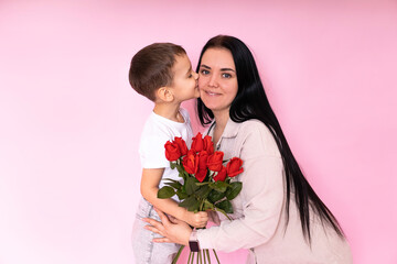 a gentle son kisses a happy mother and gives her a bouquet of roses, congratulating her on Mother's Day during the celebration of the holiday on a pink background.