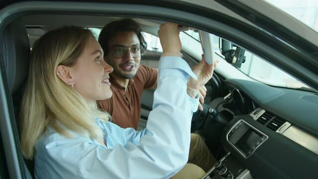 Young Caucasian woman and her Middle Eastern husband choosing new car to buy in dealership showroom sitting in one of models discussing its features