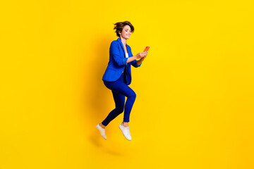 Fototapeta na wymiar Full length photo of good mood woman wear blue jacket typing samsung apple device jumping high isolated yellow color background