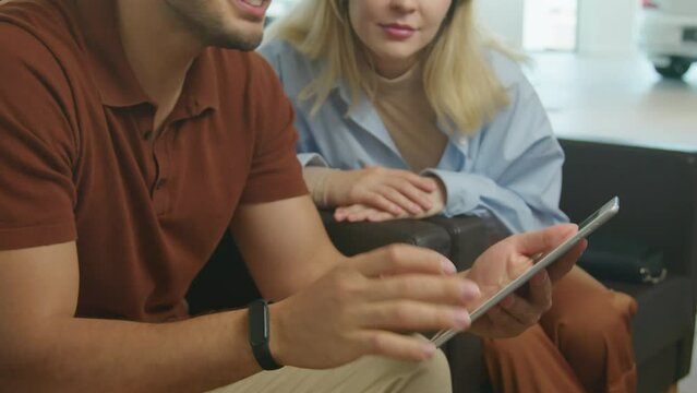 Medium section shot of young man and his wife holding digital tablet asking questions about new car model to dealership salesperson