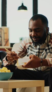 Vertical video: Happy guy eating noodles with chopsticks at home, enjoying asian food from delivery. Young adult relaxing with bottle of beer and takeaway meal, laughing at favorite tv show. Handheld