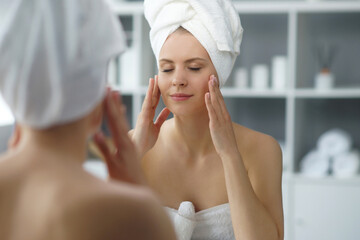 Young woman sits in the bathroom in front of the makeup mirror and does cosmetic procedures. Beautiful girl in white towel. Skin care, health, rejuvenation and spa treatment concept.