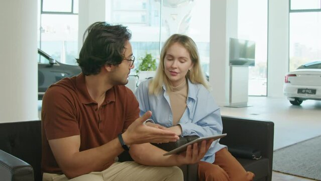 Modern young Middle Eastern man and his Caucasian wife sitting on chairs in dealership showroom holding digital tablet discussing car model features