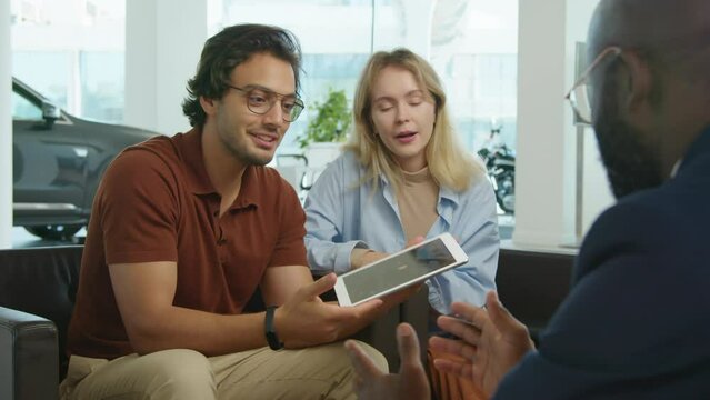 Young Middle eastern man and his Caucasian wife sitting in armchairs in front of African American dealership worker discussing car interior features