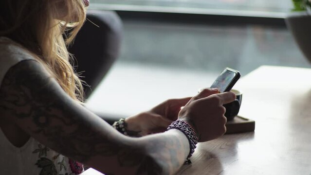 A young woman in casual attire sits by the window in a cafe, drinking coffee. Looking at the phone, typing text on social networks.