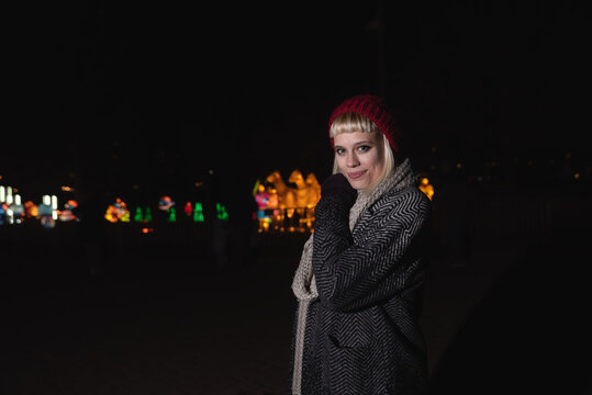 Night portrait of beautiful young fashion hipster girl with scarf and cap in coat with city street lights background