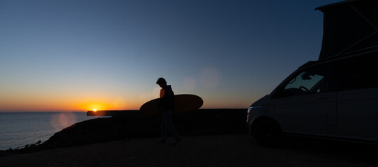 Surfer boy sitting near his mini van and looking on the ocean at summer sunset  with a surfboard on her side