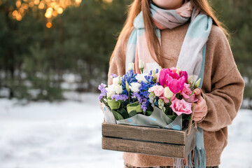 Unrecognizable caucasian teen girl with long loose hair wearing sweater and scarf holding wooden box with bright colorful spring flowers bouquet outdoors at golden hour.