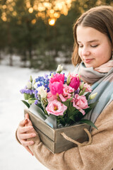 Caucasian teen girl with loose hair wearing sweater and scarf holding wooden box with bright colorful spring flowers bouquet outdoors at golden hour.