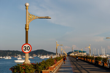 Fototapeta premium Phuket, Thailand - January 16, 2023: Chalong pier, Phuket island. A long concrete pier adorned with colorful tropical flowers and golden lanterns. Many boats, sailing and motor yachts in the sea near 