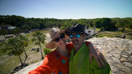 Mature woman and man couple wearing ethnic clothes, sunglasses, hats taking selfies at the top of Xcambo Mayan pyramid ruins in Mexico.