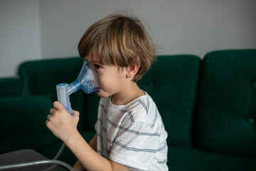 Calm, tired blonde little boy breathing with nebulizer, inhaler mask, sit on green sofa. Asthma and bronchial disease