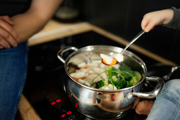 Pan with boiled vegetables and meat closeup. Baby sit near stove and try food for readiness. Healthy nutrition.