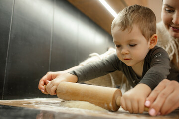Naklejka premium Happy mother with baby son roll out raw dough with wooden rolling pin on kitchen tabletop. Child help mom cooking.