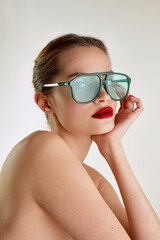Fototapeta premium Modern fashion look, attraction. Young beautiful girl with red lips posing in green sunglases over grey studio background. Concept of natural beauty, skin care, cosmetology, cosmetics, health, fashion