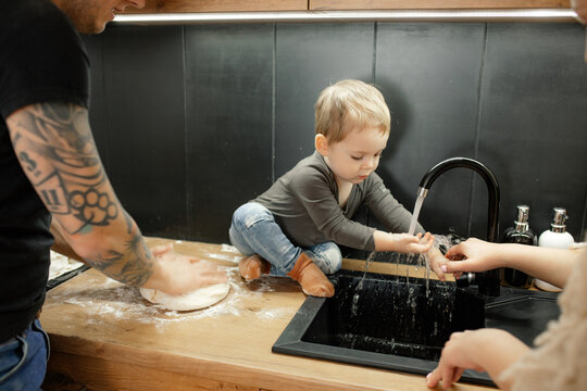 Little Child Wash Hands In Sink Sitting On Kitchen Tabletop. Baby Boy Get Dirty In Flour. Dad Knead Dough With Hands.