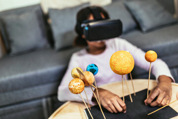 Happy african american school girl making a solar system for a school science project at home using VR glasses to immerse herself in metaverse
