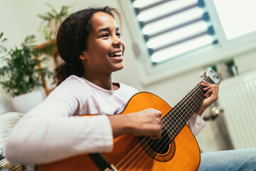 African american teenage girl sitting on couch in her room and learning to play guitar