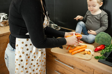 Mother with little child together cook dinner. Woman peel fresh raw carrots on wooden kitchen board. Family cooking.