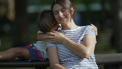 Son embracing mother sitting at park bench together. Mom and child embrace enjoying nature in beautiful sunny day