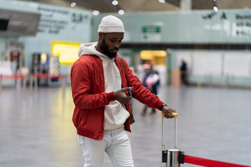 Thoughtful African American traveler man using smartphone at airport terminal, travelling with luggage bag. Passenger guy waiting her flight, checks time of flight online, standing in railway station
