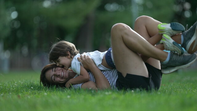 Mother and child together at park outdoors laying down in grass. Happiness motherhood concept. Son embraces parent outside kissing mom on cheek