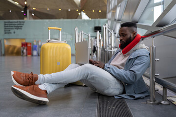 Concentrated young black man sits cross-legged waiting for flight using laptop wearing neck flight pillow with yellow suitcase communicating in social media, searching information online, typing email