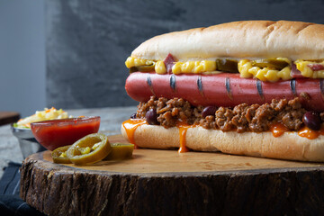 Hot dog sandwich with cheesy sauce and minced meat served with coleslaw, ketchup, mustard, and pickled pepper on a wooden tray