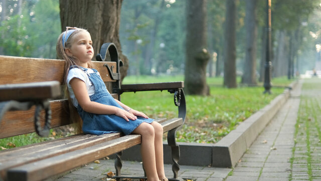 Little serious child girl sitting alone on a bench in summer park