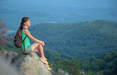 Naklejka premium Sportive female hiker sitting alone taking a break on hillside trail. Lonely woman enjoying view of evening nature from rocky cliff on wilderness path. Active lifestyle concept