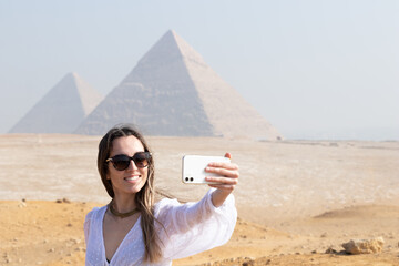 Young cheerful tourist woman taking a selfie with the Pyramids of Egypt in the blurred background
