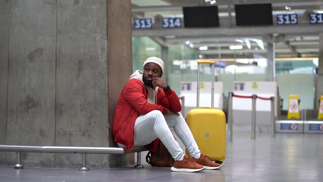 Tired African American man upset at airport his flight is delayed. Nervous traveler male waiting for a plane sitting in empty terminal with baggage. Exhausted guy on a long night connection at airport
