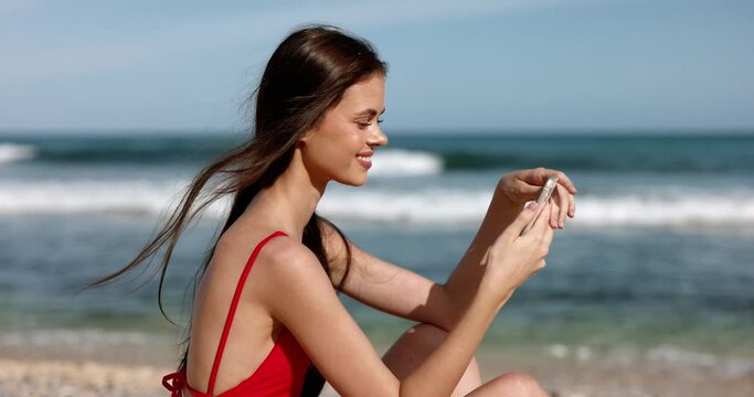 Woman sitting on the beach on the sand playing by the ocean with her phone listening to music in her headphones meditation and relaxation, beachwear red swimsuit and shorts.