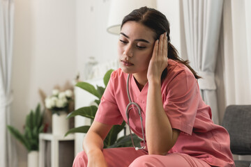 Upset female nurse sitting on a couch at home. Healthcare worker having headache. Doctors face heavy levels of stress. Young Caucasian caregiver in pink scrubs sitting hand touching temple tired face.