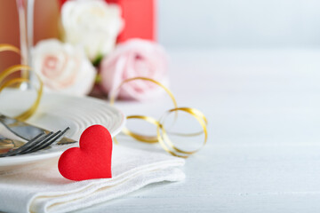 Valentines Day or Romantic dinner concept. Romantic table setting, silverware,  wine glasses, gift box, roses and symbol of love red heart on white background. Romantic Dinner Valentines Day concept.