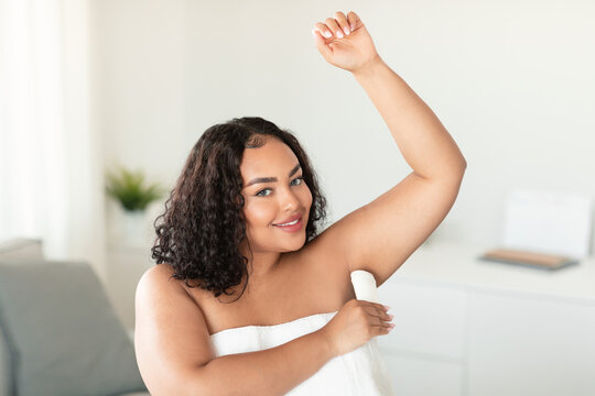 Positive black overweight woman using antiperspirant stick for underarms, standing wrapped in towel at home