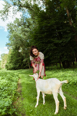 Vertical image woman dressed traditional fashionable ukrainian embroidery vyshyvanka dress shirt ethnic costume cloth feeding goat with leaves grass in the forest village. Ukrainian culture

