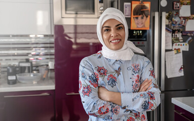Middle-aged muslim woman smiles poses in Kitchen wearing hijab headscarf. Islamic female does...