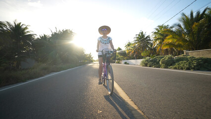 Pretty mature senior woman holding her hat while biking into the sun wearing sunglasses in a tropical setting.