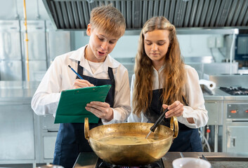 Teenage boys and girls wearing aprons and concentrating on cooking class are cooking soup and writing down baking recipes together.